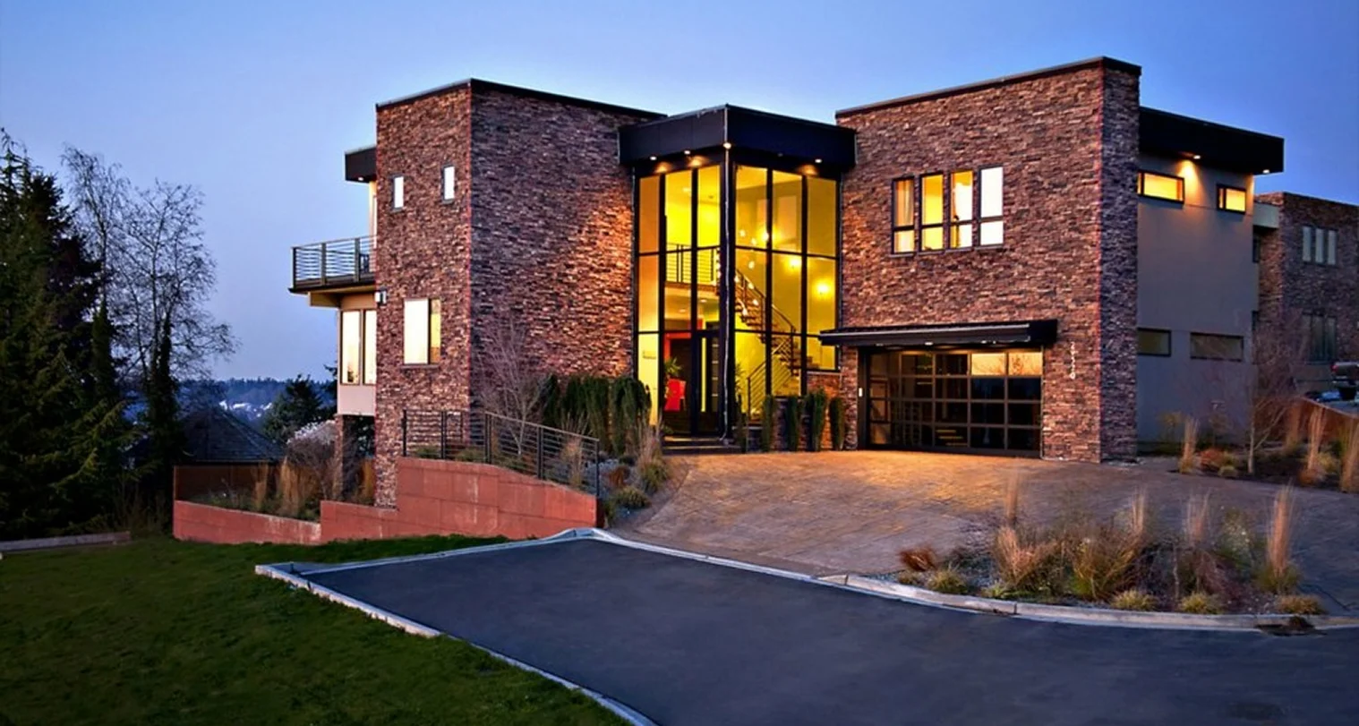 A modern house with a stone exterior and tall glass entryway glowing in the evening light.